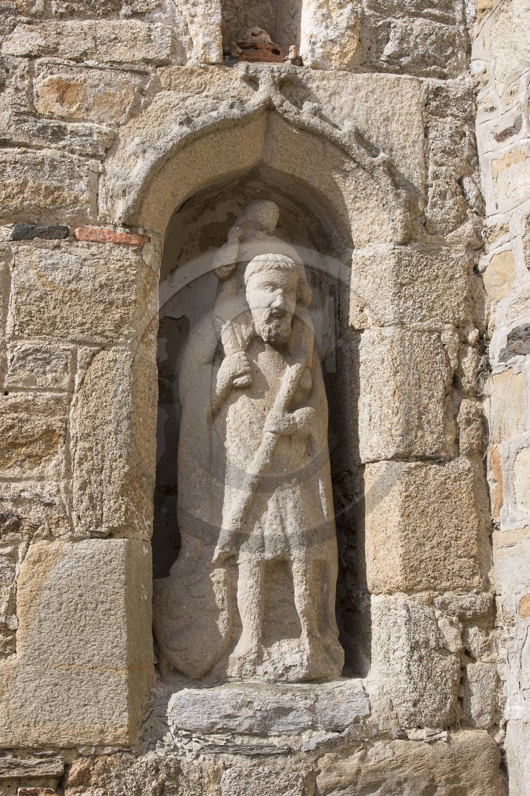 La statue de Saint-Christophe à Saint-Jacut de la Mer ( 22 ). Statue monolithe en granite abritée sous une niche aménagée dans le mur antérieur d'une maison, représentant saint Christophe portant l'enfant Jésus sur ses épaules, tenant dans sa main droite le pied du Christ et dans sa main gauche un bâton feuillu dont la partie basse est accompagnée d'un écu scutiforme armorié (probablement trois merlettes).